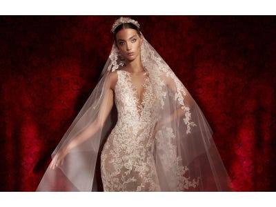 Braunhaarige Braut mit durchsichtigem weißen Brautkleid aus Spitze und langem Spitzenschleier vor einer Wand aus roten Rosen