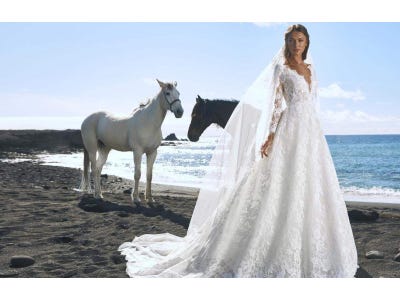 Modella sulla spiaggia che indossa un abito da sposa di pizzo con maniche lunghe e scollo a V e un lungo velo
