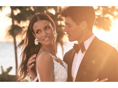 Noiva com brincos e um vestido de bordados florais abraça noivo ao pôr do sol  