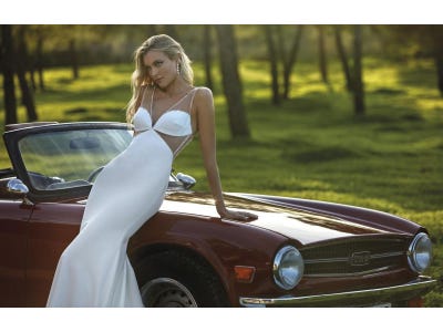 Mujer vestida de novia con un vestido moderno apoyada sobre un coche rojo