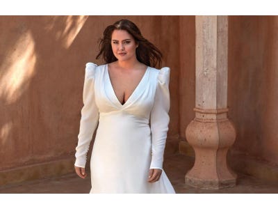 Curvy brunette woman in an A-line crepe wedding gown featuring a V-neck with long sleeves and puff shoulders standing outisde