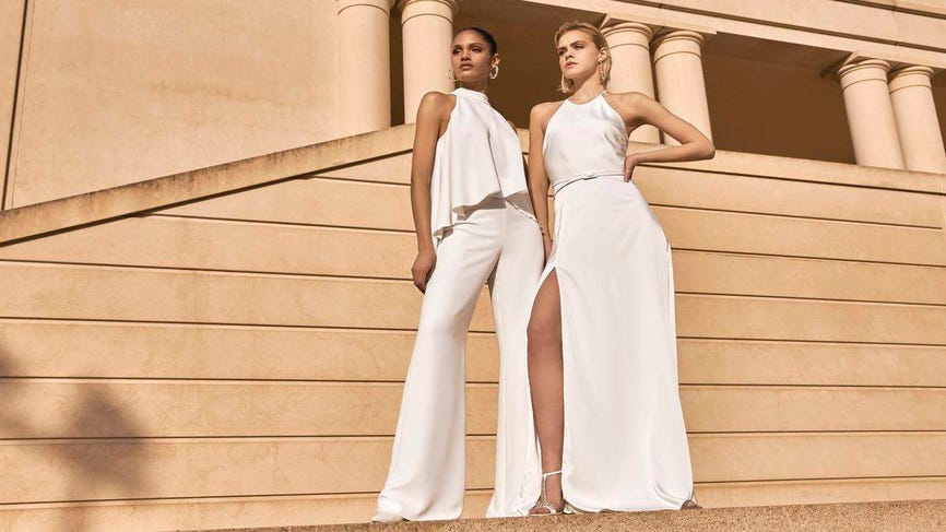 Due donne vestite, una con un vestito bianco a due pezzi e l'altra con un vestito morbido bianco davanti ad un monumento. 