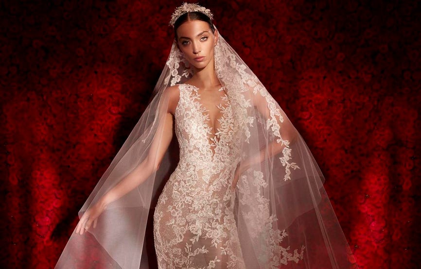 Braunhaarige Braut mit durchsichtigem weißen Brautkleid aus Spitze und langem Spitzenschleier vor einer Wand aus roten Rosen