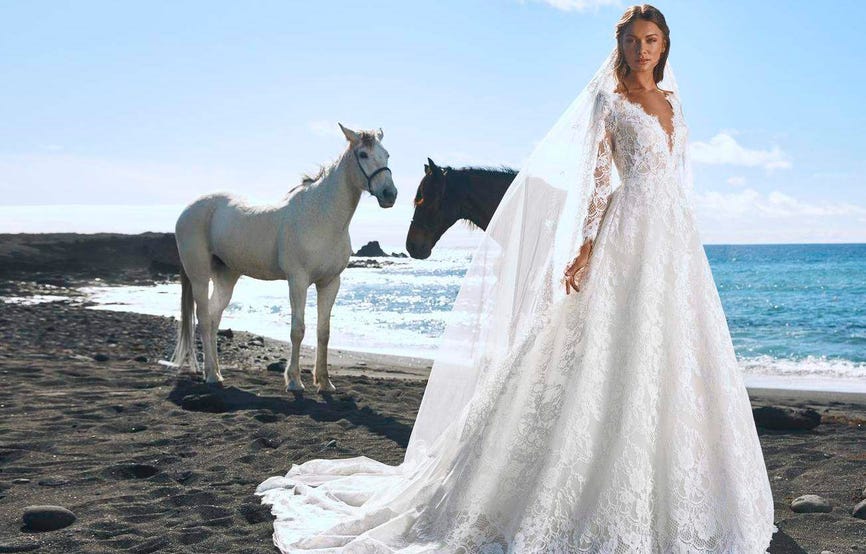 Modella sulla spiaggia che indossa un abito da sposa di pizzo con maniche lunghe e scollo a V e un lungo velo