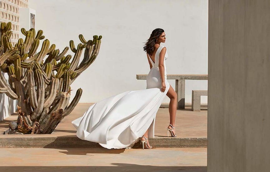 Une élégante mariée en robe de mariée blanche et talons se tient sur une corniche, rayonnante de bonheur et de grâce.