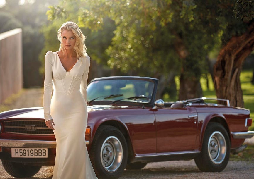 Femme dans une robe de mariée col V à manches longues, se tenant devant une voiture de collection