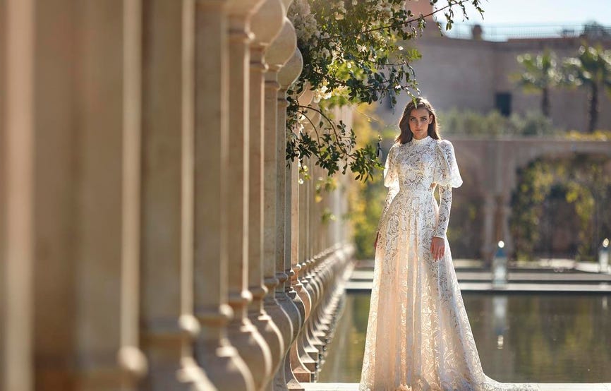 Woman in a puffy long-sleeved wedding dress with intricate lace detailing standing next to water