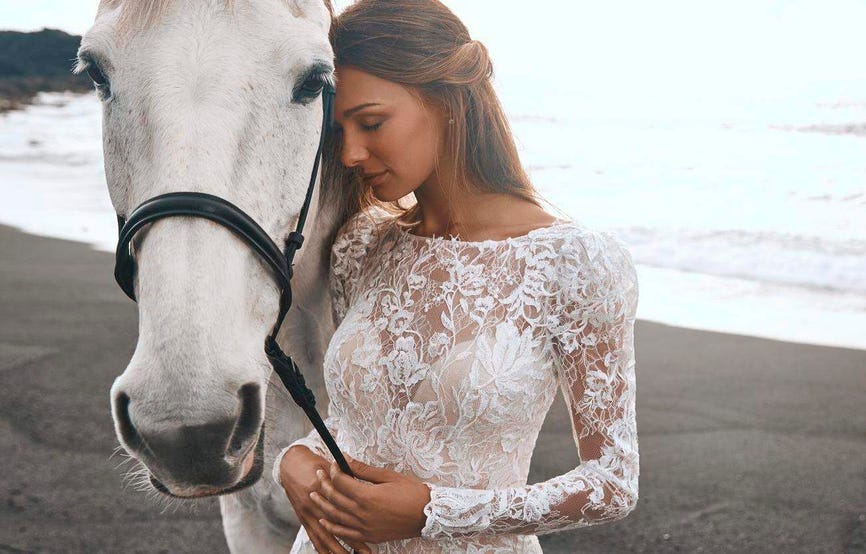 Ρομαντική νύφη που φοράει δαντελένιο νυφικό με μακρύ μανίκι κρατώντας ένα άλογο στην παραλία