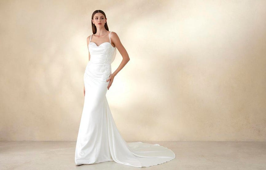 Brunette wearing a long mermaid-cut wedding dress, made with satin, posing in an empty room