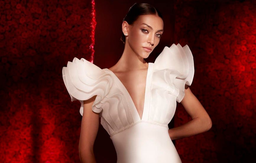 Brunette in a bun wearing a mermaid crepe bridal gown with statement bodice ruffles standing against a red background