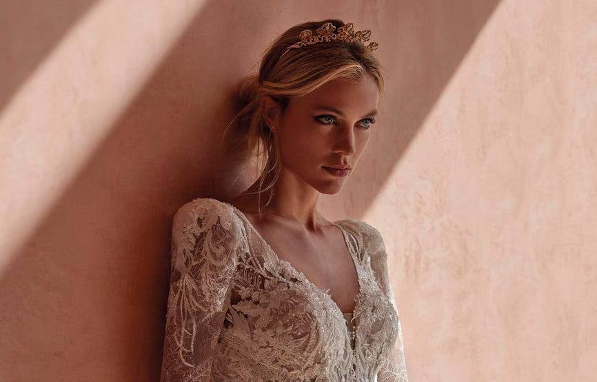 Blonde wearing a mermaid wedding gown with lace detailing and deep V-neck, standing against a wall
