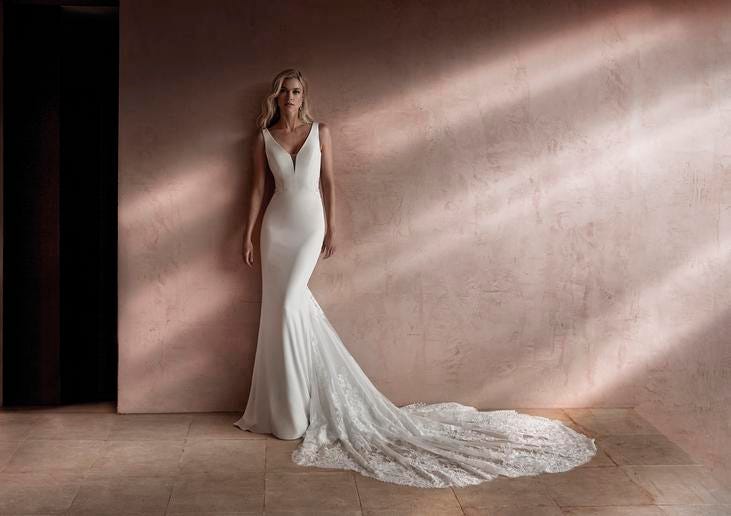 Blond kobieta stojąca przy ścianie w długiej prostej skromnej sukni ślubnej z głębokim dekoltem i delikatnym trenem
