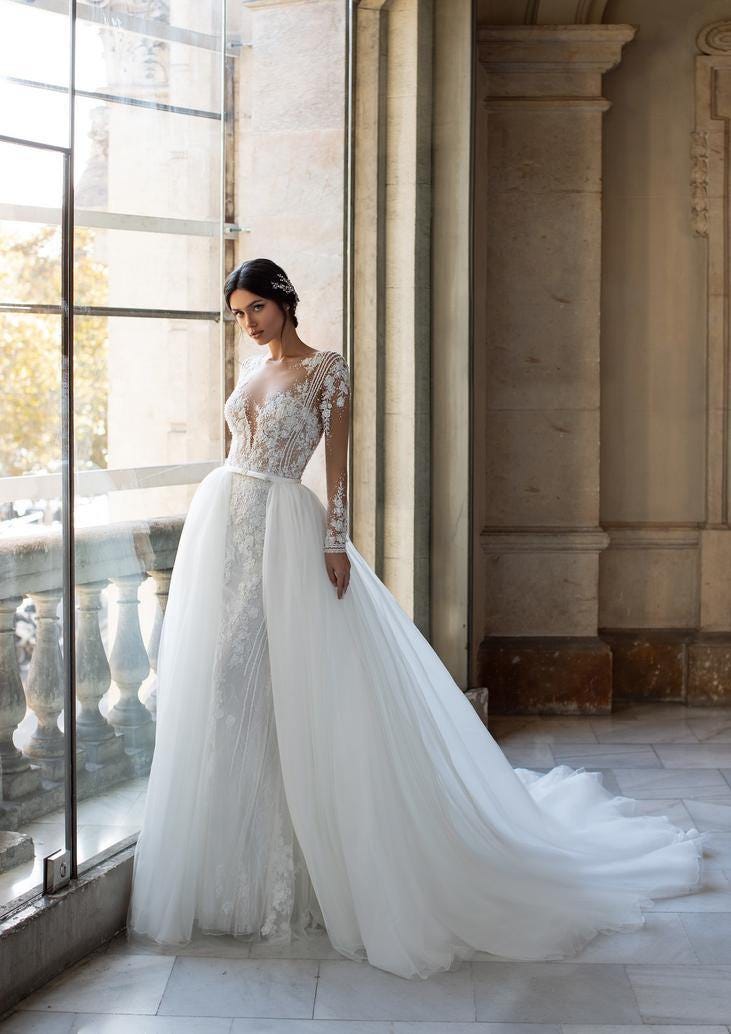 Une mariée, habillée d'une robe de mariée en dentelle à manches longues, se tient à côté d'une fenêtre, rayonnante de beauté.