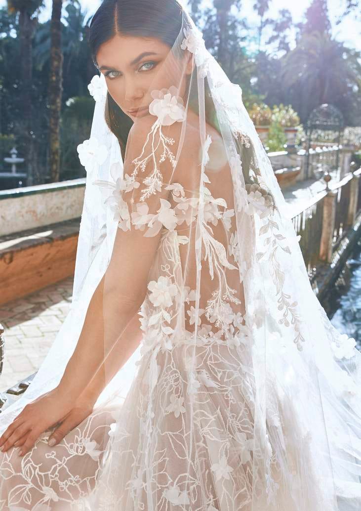 Noiva de cabelo longo a usar um vestido com decote cai cai e um véu adornado com bordados de flores