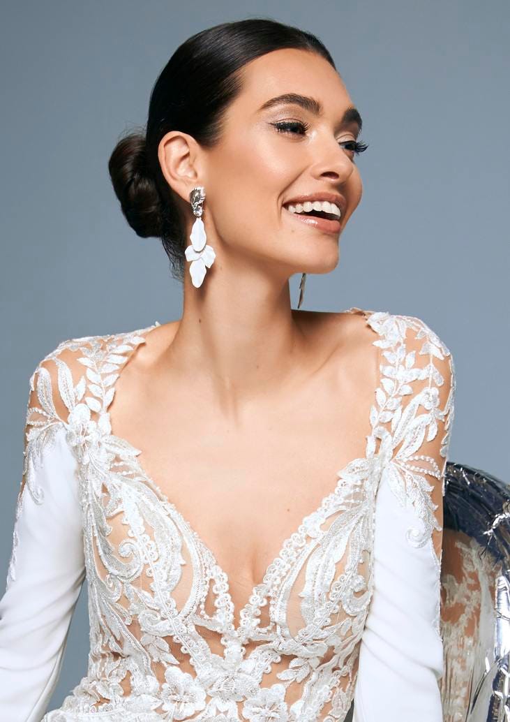 Noiva posando de perfil com um vestido de mangas longas combinado com um coque baixo e uns brincos brancos longos