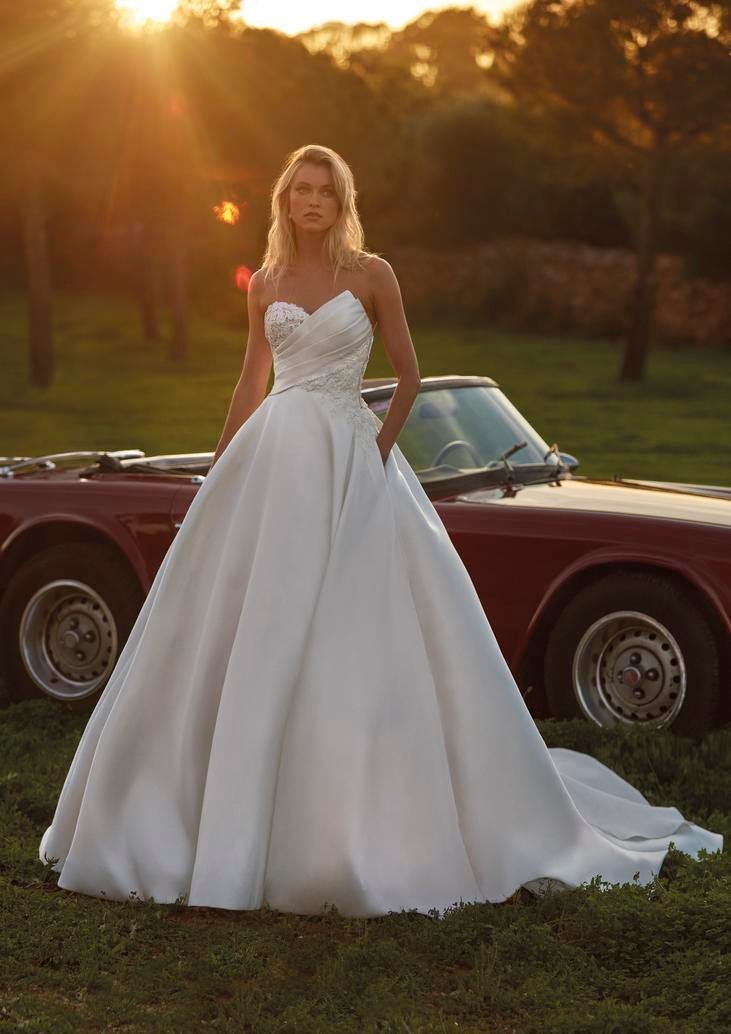 Mariée vêtue d'une robe de mariée princesse sans bretelles avec jupe bouffante, se tient devant une vieille voiture de luxe.