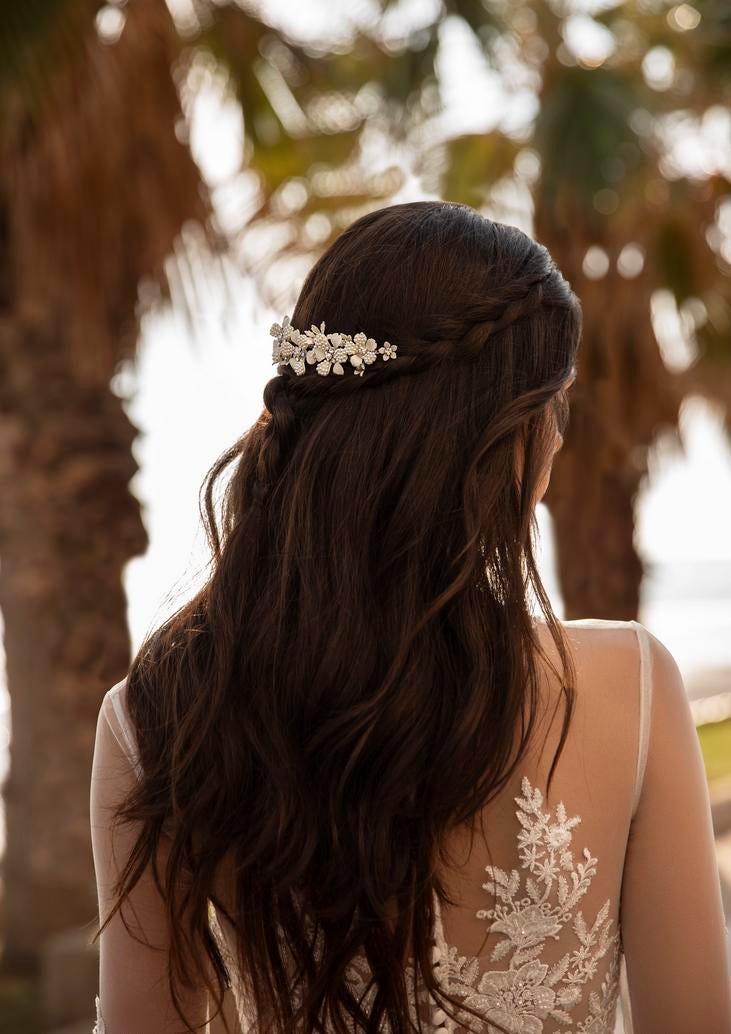 A bride from behind with a floral hairpiece in her braided dark hair, and a lace-detail wedding dress, against a palm tree backdrop. 