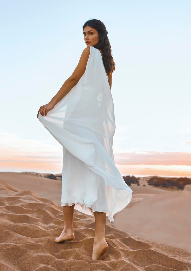 Dunkelhaarige Frau in weißem Kleid aus leichtem Stoff von hinten über die Schulter blickend im Sand
