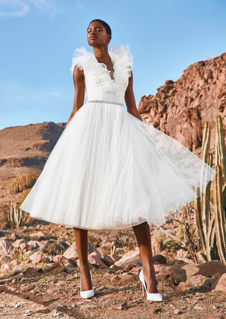 Czarnoskóra kobieta stojąca na skalistym podłożu prezentująca krótką tiulową suknię ślubną i białe buty na obcasie