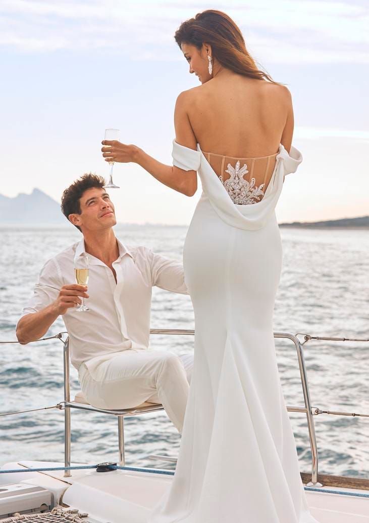 Woman wearing wedding dress with a scoop back facing a man in a chair with a glass of champagne by the ocean