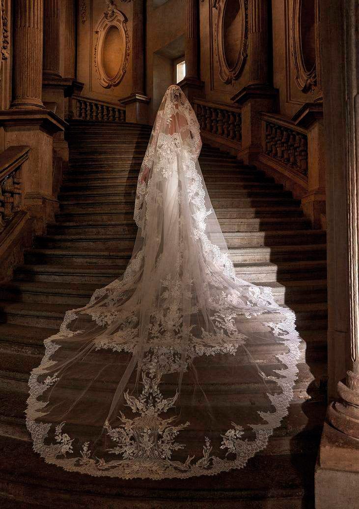 Donna di spalle su lunga scalinata che indossa un abito da sposa principesco con lungo velo di pizzo.
