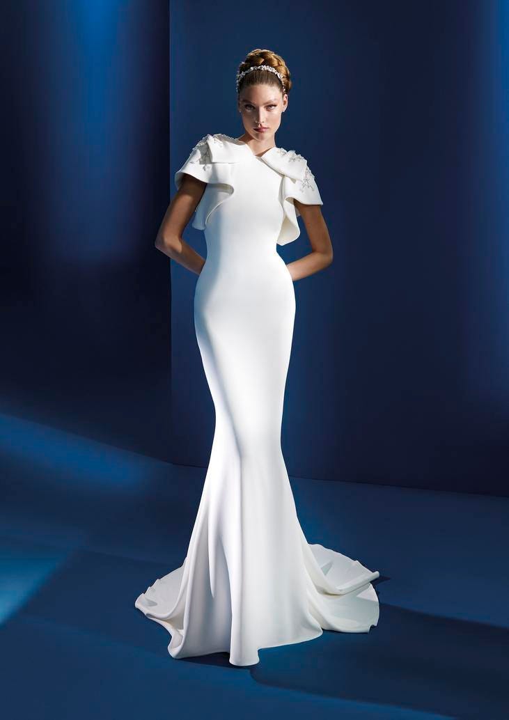 Blonde in a crepe wedding dress with bateau neckline and ruffle-effect sleeves, standing against a blue background