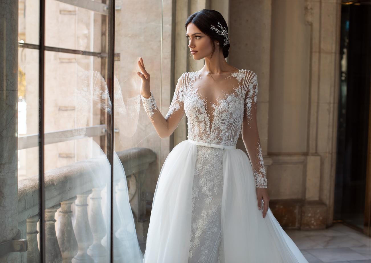 Dunkelhaarige Braut in weißem Brautkleid aus Spitze und Glitzer und Tüllbrautgürtel vor einer Glaswand in einem Schloss 