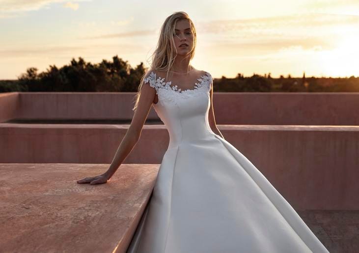 Blonde-haired woman wearing a scoop-neck wedding dress with short sleeves and floral detailing lining the neckline. 
