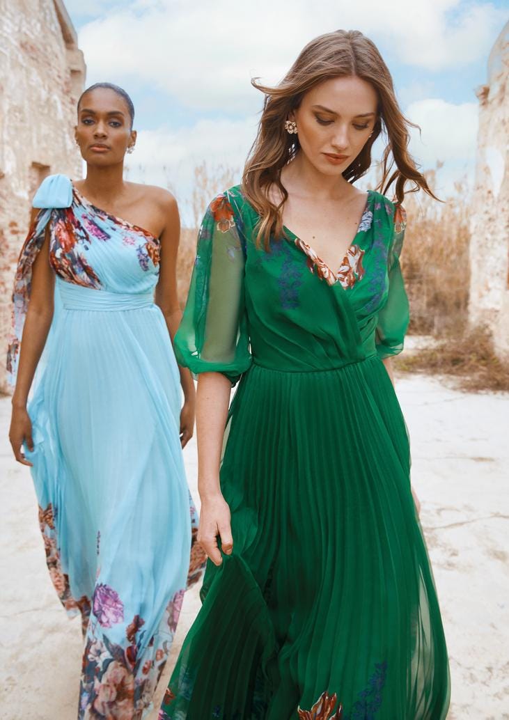Frau in hellblauem langen schulterfreien Kleid mit bunten Blumenmustern neben Frau in grünem langen Kleid mit halblangen Ärmeln und bunten Blumenmustern 