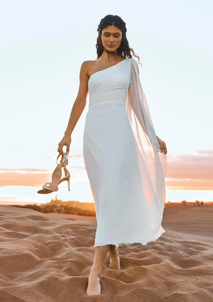 Brunette in a tea-length sheath wedding dress with a chiffon shoulder accent, walking on the beach and holding high heels.