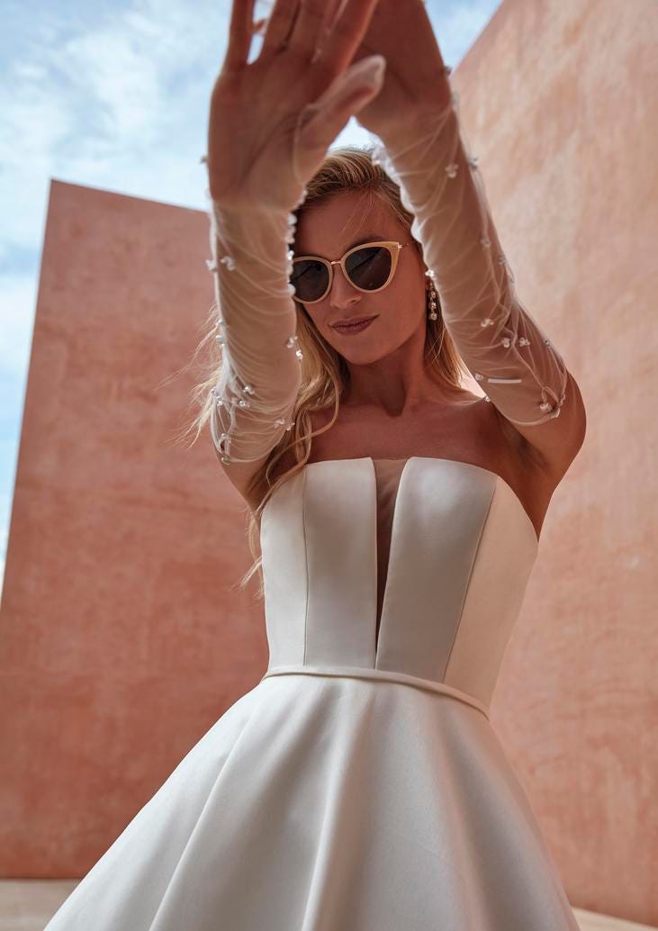 Blonde Frau in schulterfreiem ALinien Brautkleid mit druchsichtigen Armstulpen und Sonnenbrille die Arme verkreuzend