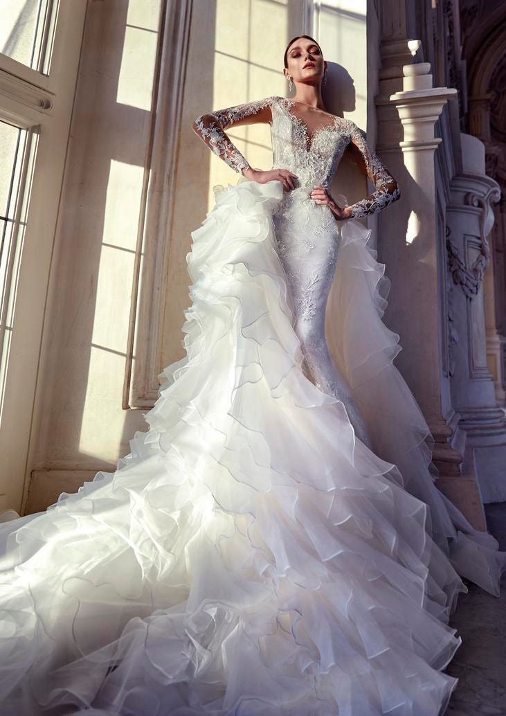 Modelo posa com vestido de noiva sereia com cauda longa de babados, com decote coração, mangas longas bordadas e penteado preso