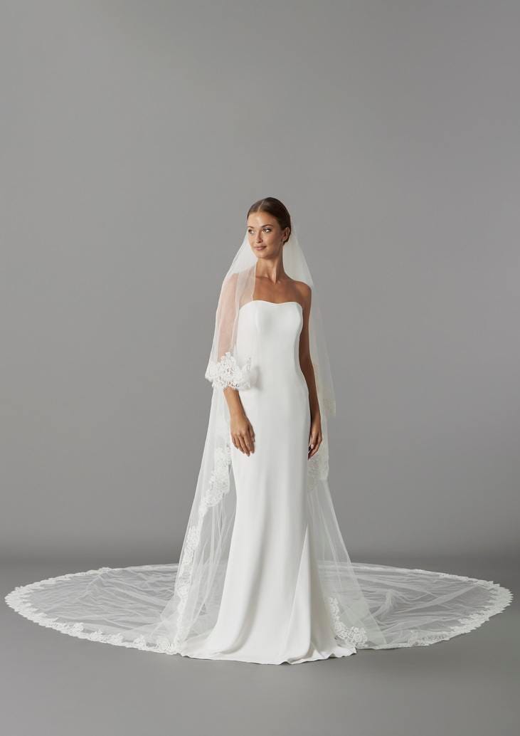 Modella che indossa un abito da sposa semplice e un velo da sposa di tulle corto davanti e lungo dietro ricamato sui bordi.