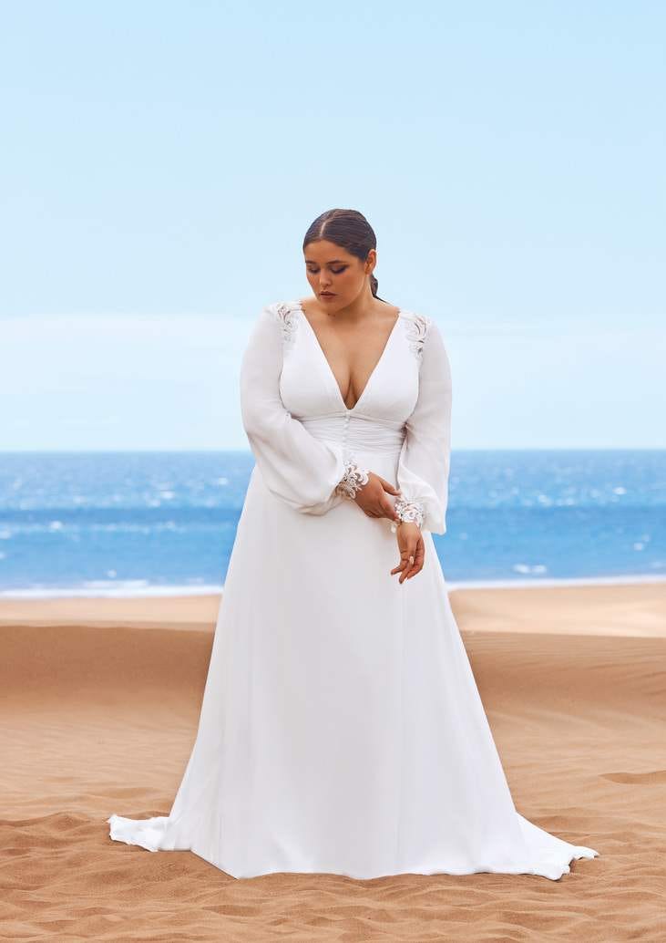 Modella sulla spiaggia che indossa un abito da sposa plus size con maniche lunche e scollatura a V.