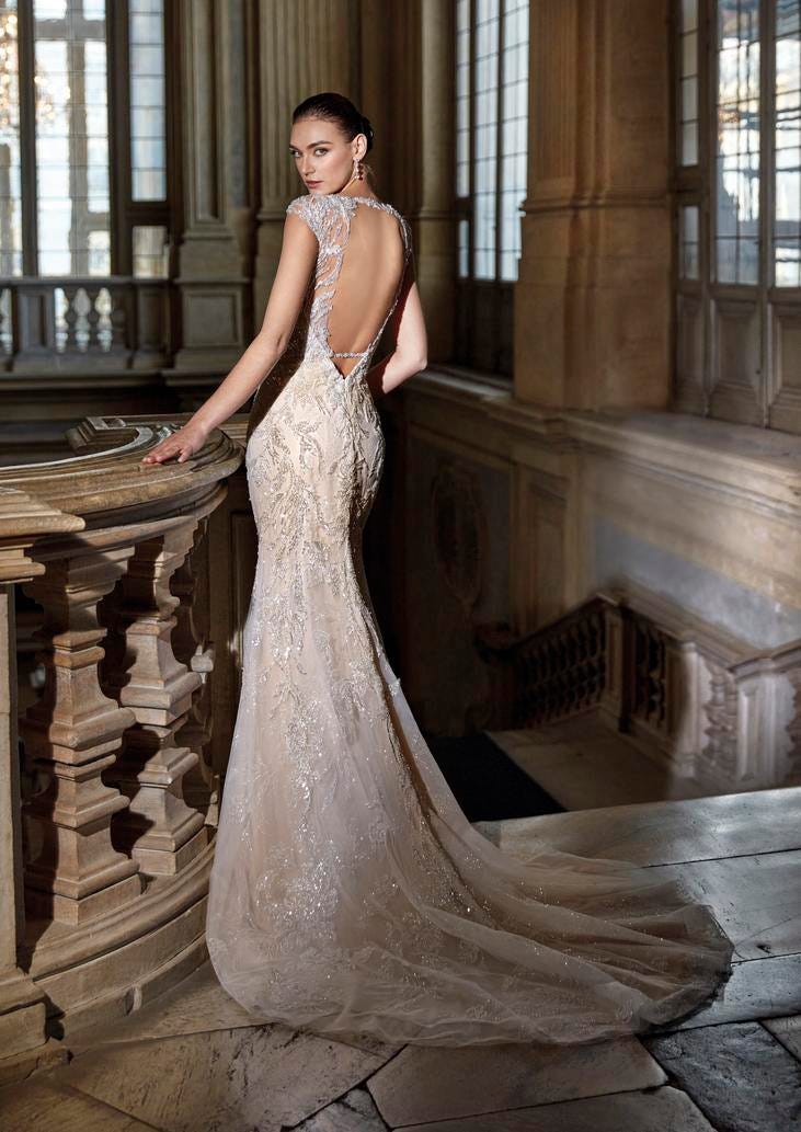Woman wearing a glitter mermaid wedding dress with an open back and metallic embriodery, standing near a staircase