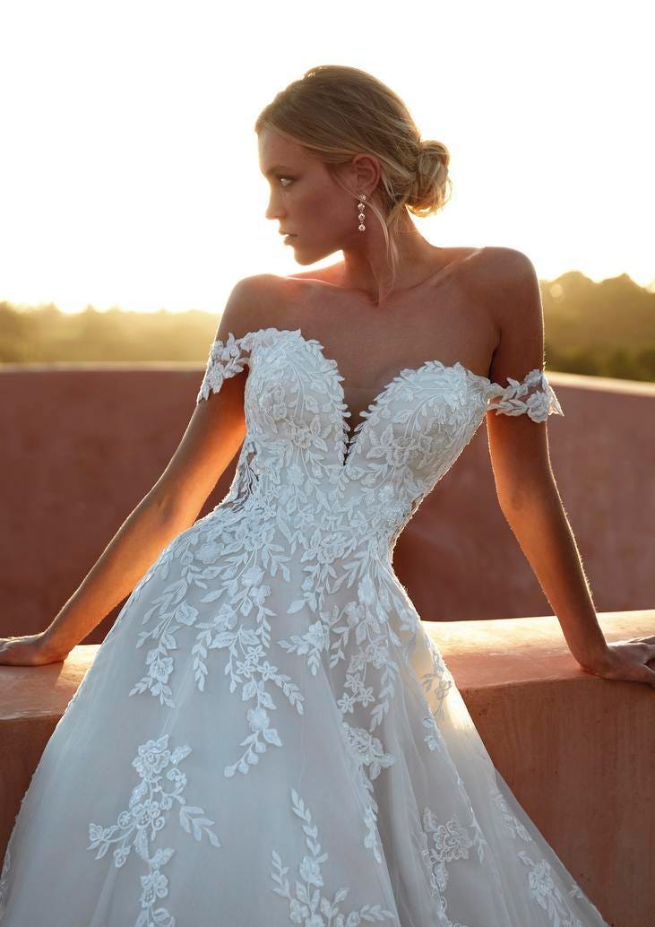 Blonde wearing a bun in an A-line tulle wedding dress with floral lace applique, standing outside and looking to the side