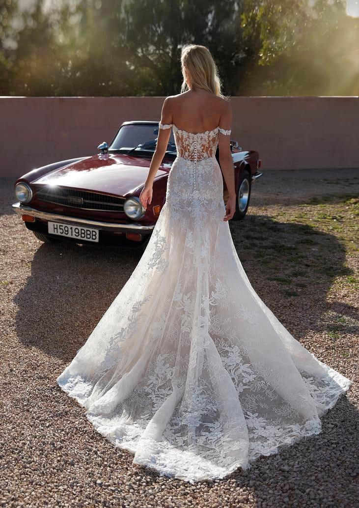 Blonde wearing an off-the-shoulder mermaid wedding dress and sculpurala seam at the back, walking towards a car outside