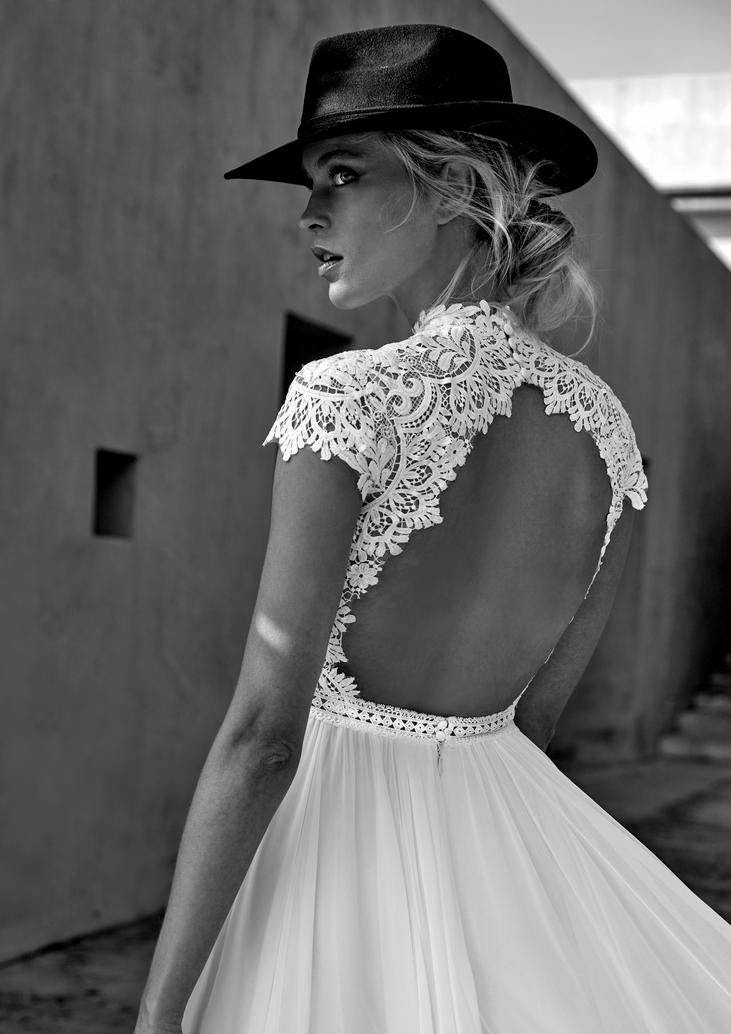 Frau mit Cowboyhut trägt ein weißes Brautkleid mit Rückenausschnitt Ein Schwarz-Weiß Foto von einer Frau mit hochgestreckten, blonden Haaren schaut über ihre Schulter und trägt einen Cowboyhut und ein Brautkleid mit detailverziertem Rückenausschnitt.