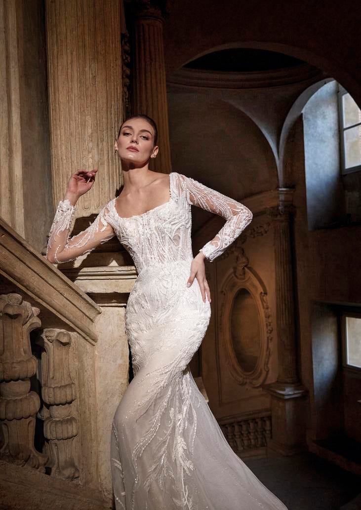 Woman wearing a mermaid statement wedding gown with square neckline and rich lace embroidery, standing at a staircase
