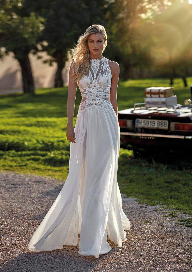 Blonde woman wearing a flowing chiffon wedding dress, standing beside a car, surrounded by greenery