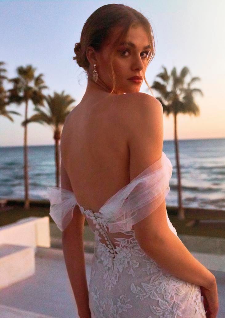 Woman wearing a mermaid wedding dress with exposed back, standing near a beach with palm trees