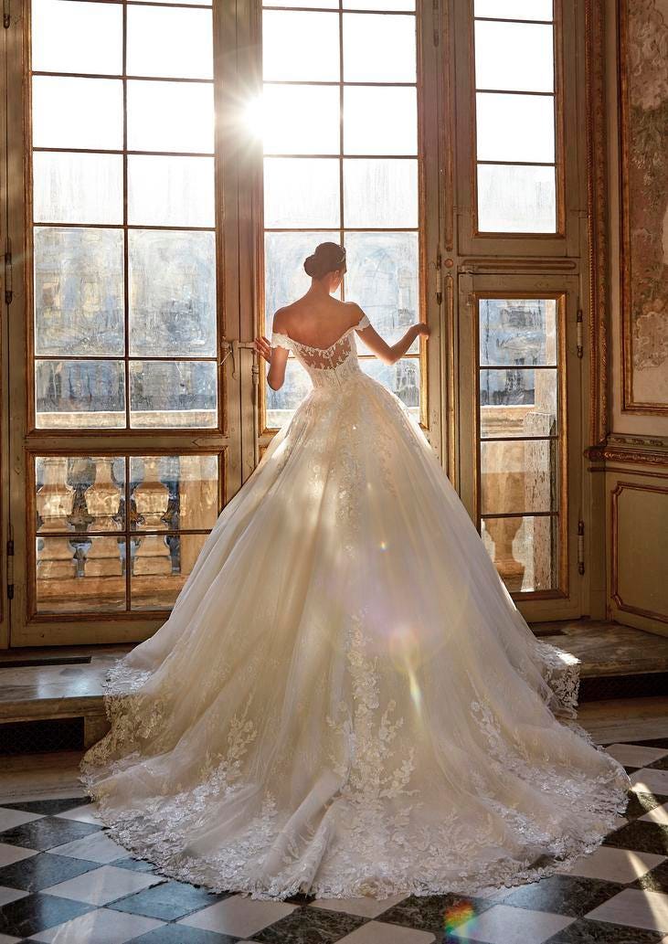 Woman wearing a sleeveless princess wedding dress with illusion back and long train, looking outside the window in a ballroom