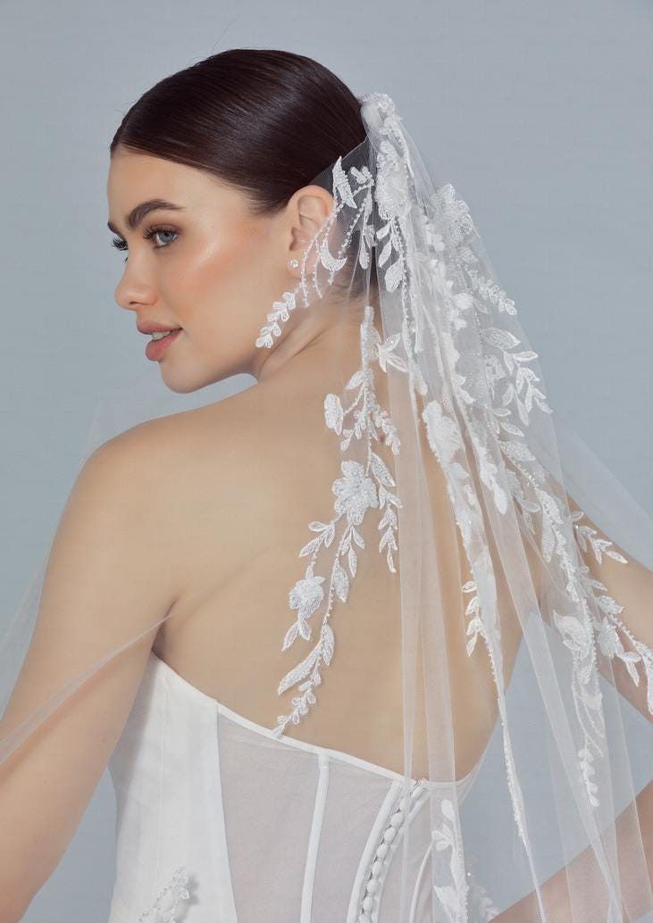 Brunette wearing a strapless bridal gown and an off-white wedding veil with intricate lace, looking from behind.